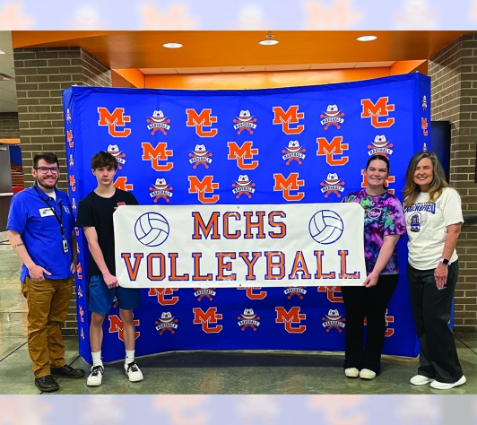 Nick Adams of the library stands in front of a MC backdrop with two students and a teacher. The students are holding a vinyl MCHS Volleyball sign made in the library's MakerSpace.