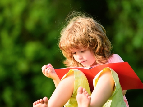 A preschool age child sitting in green grass reading a red book