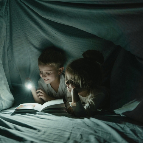 two children in a pillow fort reading a book with a flashlight