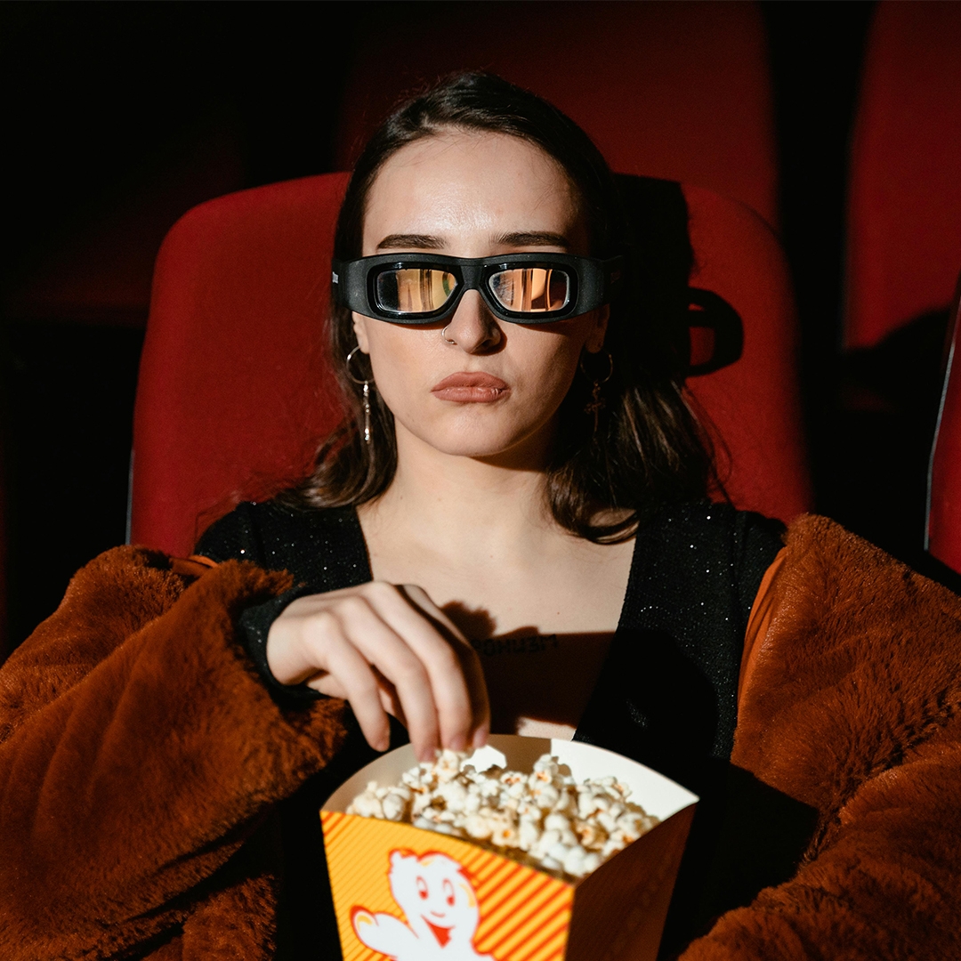 a girl with nose rings, a fur jacket, and weird glasses watching a movie and eating popcorn